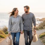 Un couple souriant marche côte à côte avec de petits sacs de voyage sur un sentier côtier paisible, profitant d’un week-end romantique.