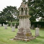 Ancien calvaire breton en pierre dressé au bord d’un cimetière communal, éclairé par une lumière douce et diffuse.